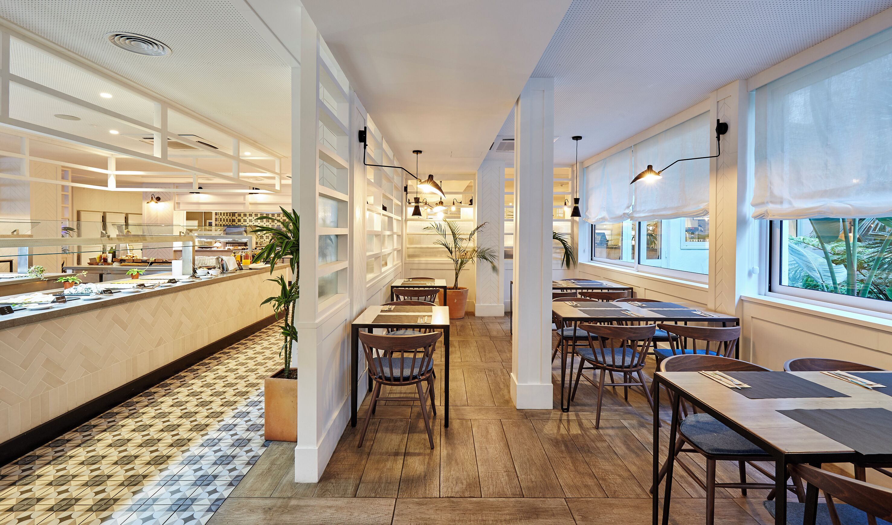 Un restaurant moderne avec un long comptoir, des tables et des chaises en bois. On y trouve des plantes en pot et une vue sur la verdure à l’extérieur.