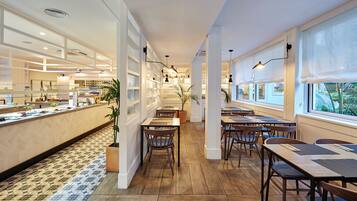 Un restaurante moderno con una barra larga, mesas y sillas de madera. Hay plantas en macetas y vistas al exterior con vegetación.