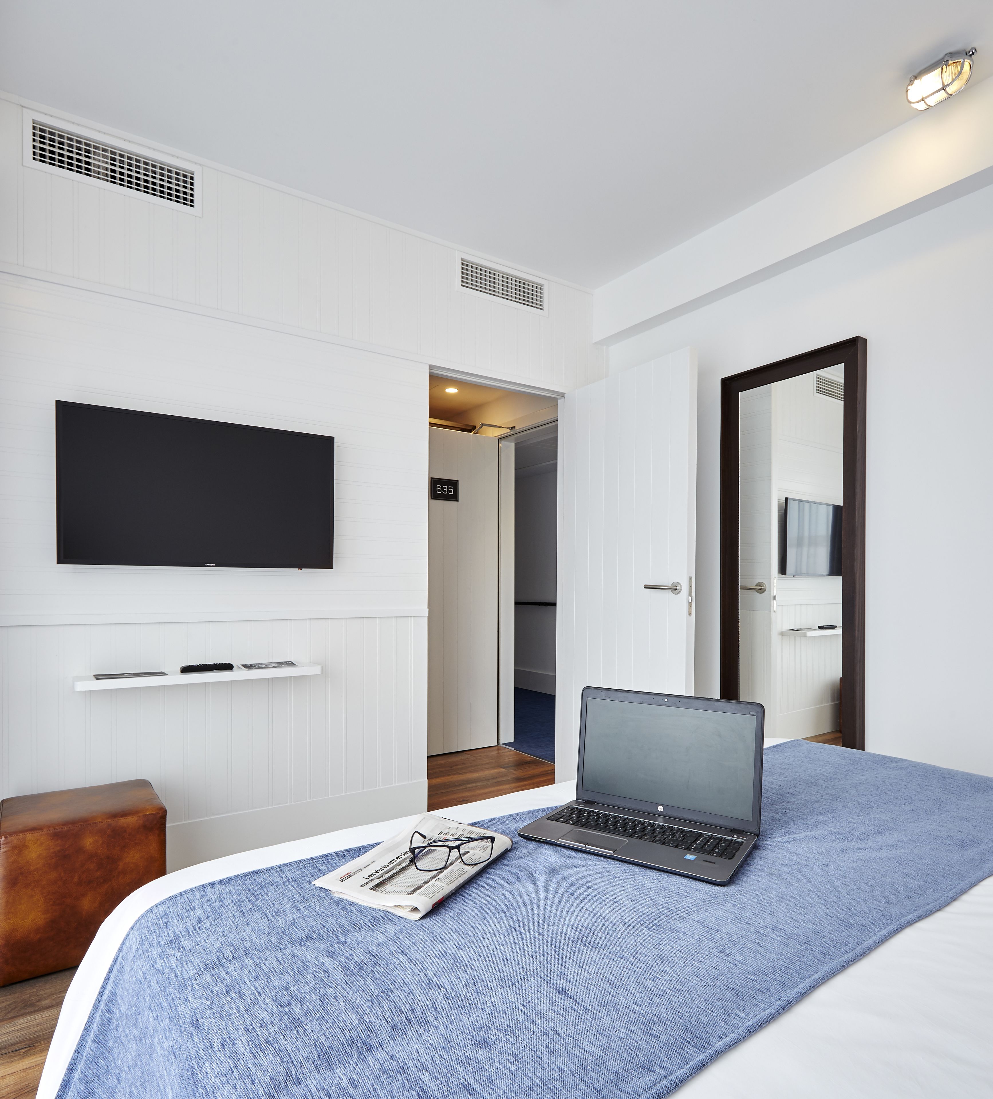 Una habitación de hotel moderna con un televisor de pantalla plana montado en la pared, una computadora portátil sobre la cama y un banco de madera.