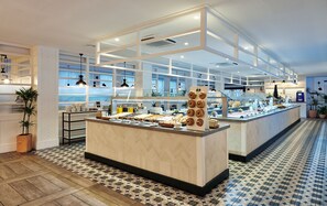 Un área de bufé con diversas opciones de comida, un mostrador con pastelería y un suelo de baldosas con diseño geométrico.