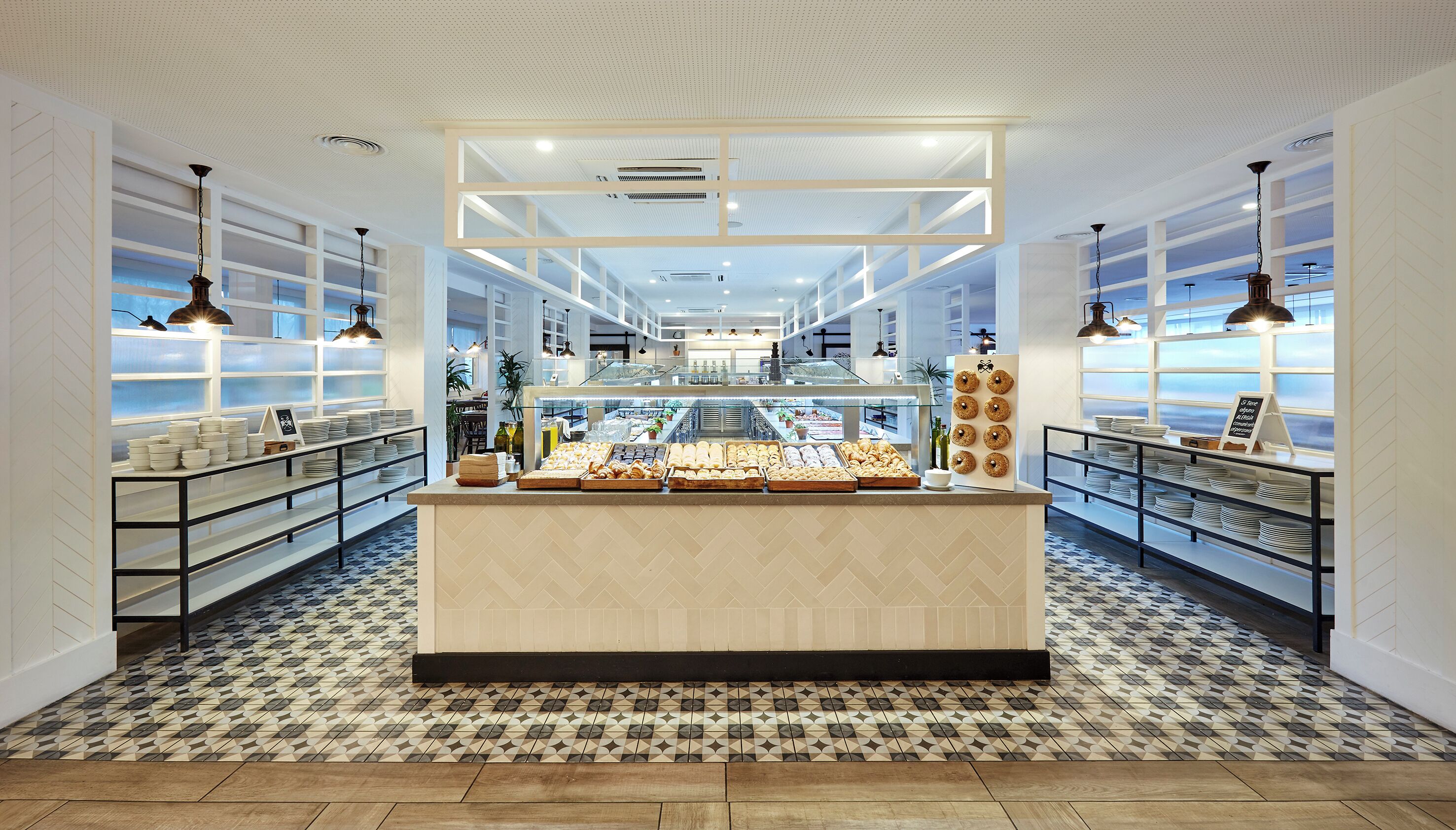 A bakery counter with pastries and bread, surrounded by shelves of plates and bowls, in a bright, modern interior with patterned flooring.