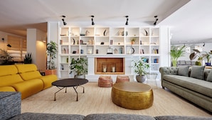 Una sala moderna con un sofá amarillo, un sofá gris y una mesa de centro redonda dorada. Hay varias estanterías con libros y objetos decorativos, y una chimenea en el centro.