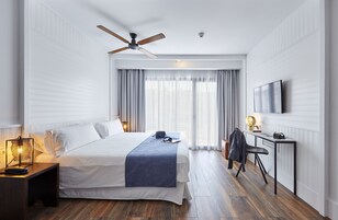 A modern hotel room with a large bed, a desk, a chair, a ceiling fan, and a TV mounted on the wall.