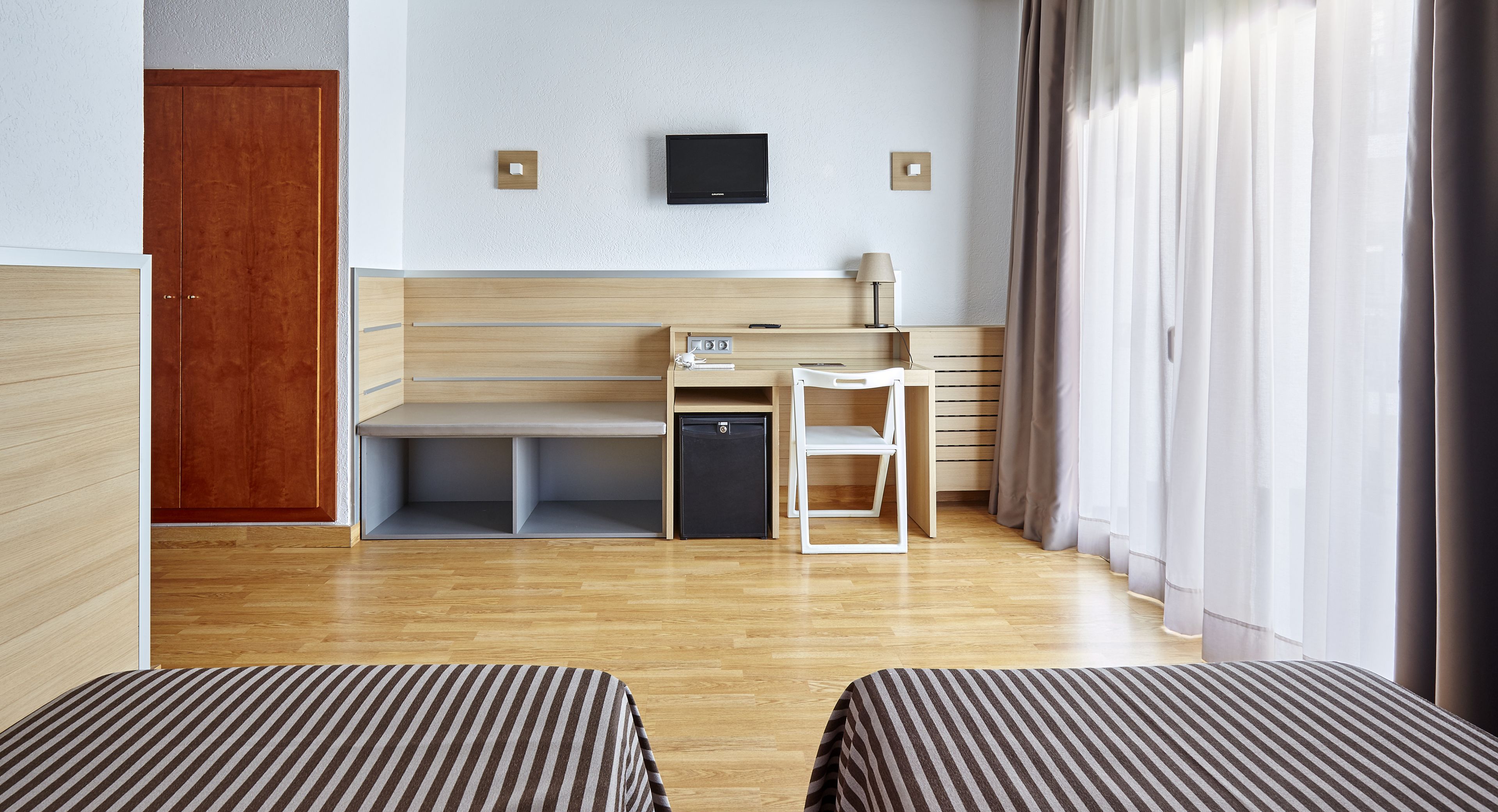 Una habitación de hotel con suelo de madera, una cama con ropa de cama a rayas, un escritorio con lámpara, un televisor en la pared y un armario.