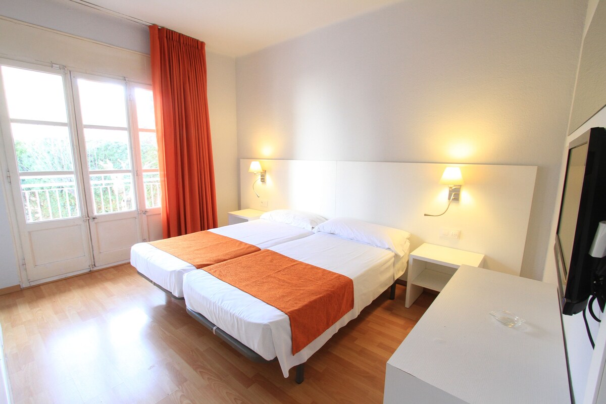 Un lit double avec un couvre-lit orange, deux tables de chevet, un bureau blanc et une grande fenêtre avec des rideaux.