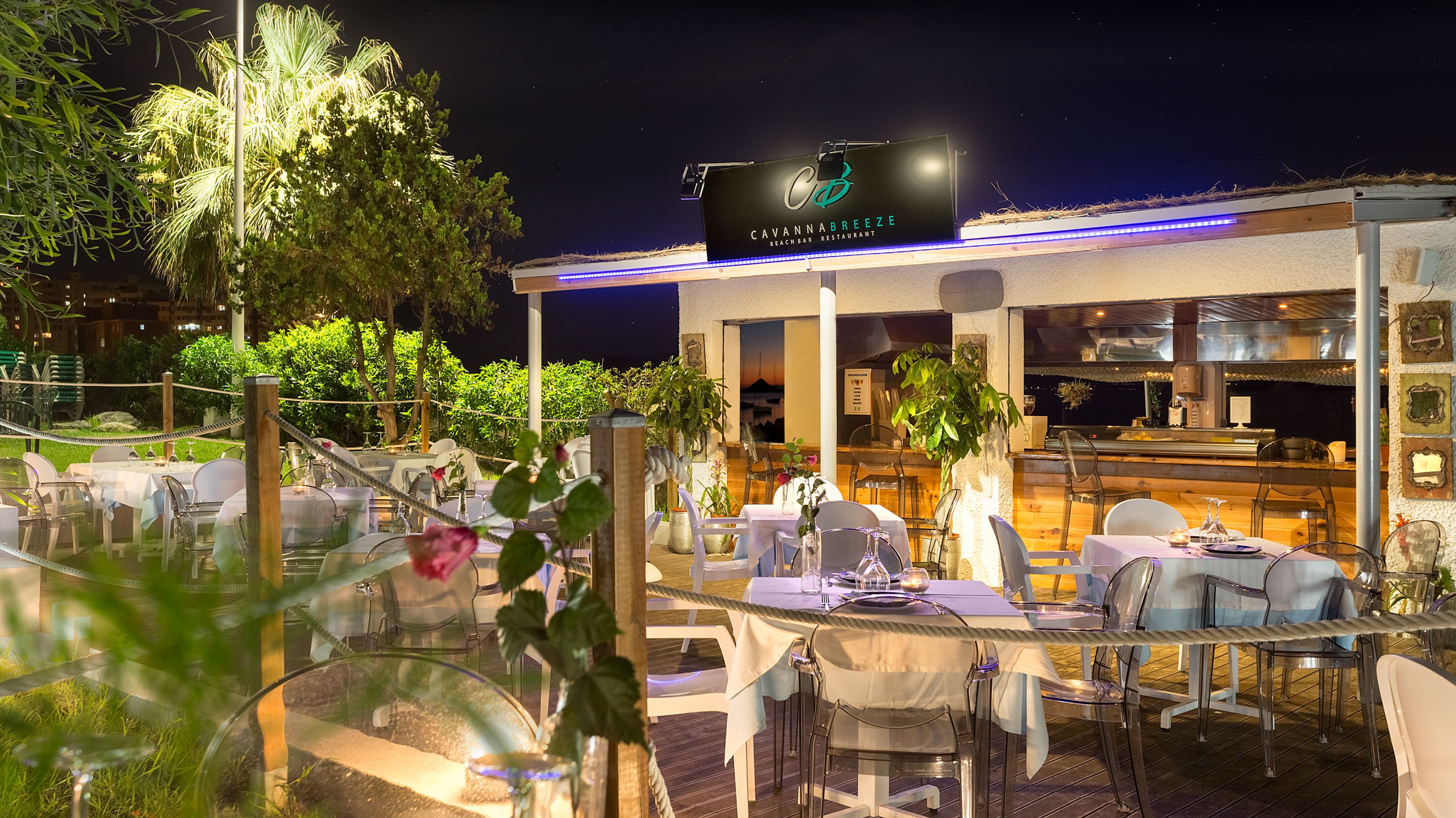 Un restaurante al aire libre con mesas y sillas, un letrero que dice Cavannabrezz Beach Bar Restaurant y vistas al cielo nocturno.