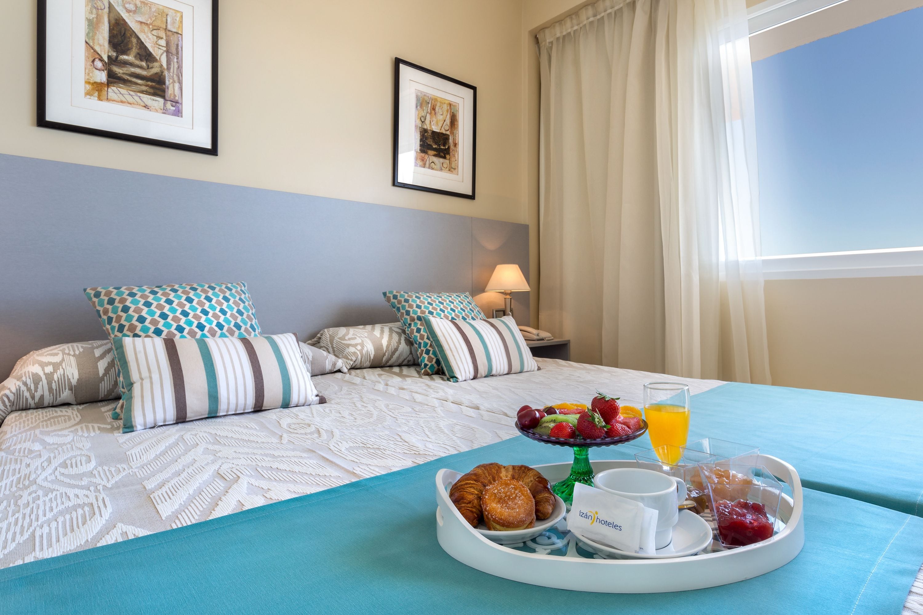A hotel room with a bed, a breakfast tray with croissants, fruit, and juice, and two framed pictures on the wall.