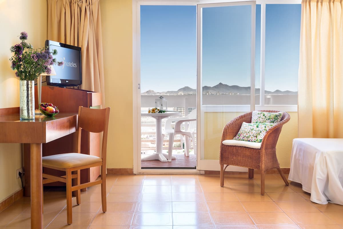 Ein Hotelzimmer mit Fernseher, einem Esstisch mit Stühlen, einem Korbsessel mit Kissen, einem Bett und einem Balkon mit Bergblick.