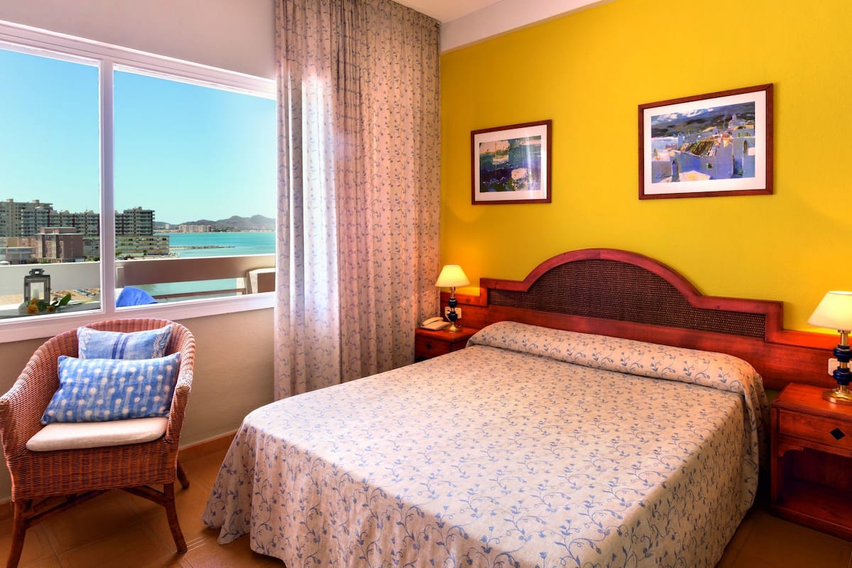 Ein Hotelzimmer mit einem Bett, zwei Nachttischlampen, einem Korbstuhl, einem Fenster mit Blick auf die Stadt und zwei gerahmten Bildern an der Wand.