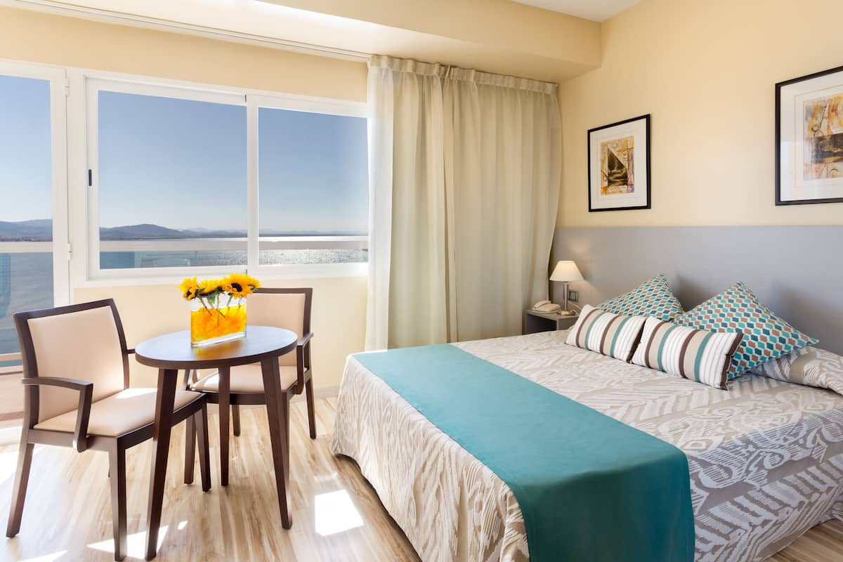 Ein Hotelzimmer mit einem Bett, einem Tisch mit Stühlen, einer Blumenvase, einem Fenster mit Meerblick und zwei gerahmten Bildern an der Wand.
