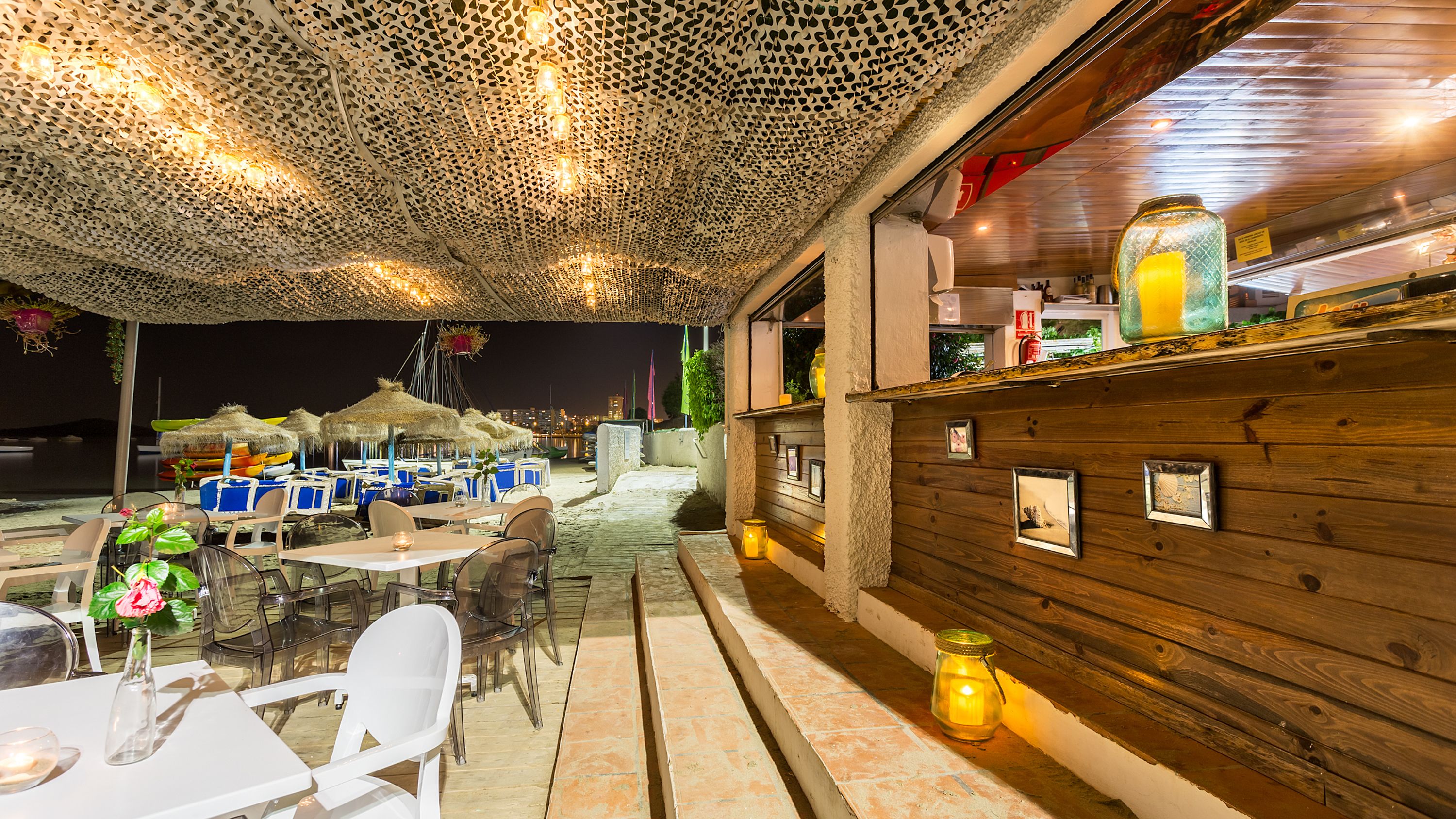Un área de comedor al aire libre con mesas y sillas, una pared de madera con cuadros enmarcados y un techo con un diseño perforado original.
