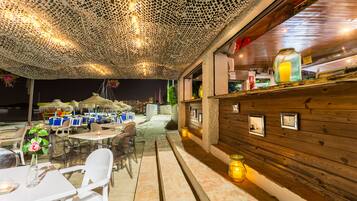 Un área de comedor al aire libre con mesas y sillas, una pared de madera con cuadros enmarcados y un techo con un diseño perforado original.
