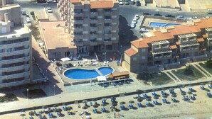A high-rise hotel with multiple balconies, a swimming pool, and a beach area with sun loungers and umbrellas.