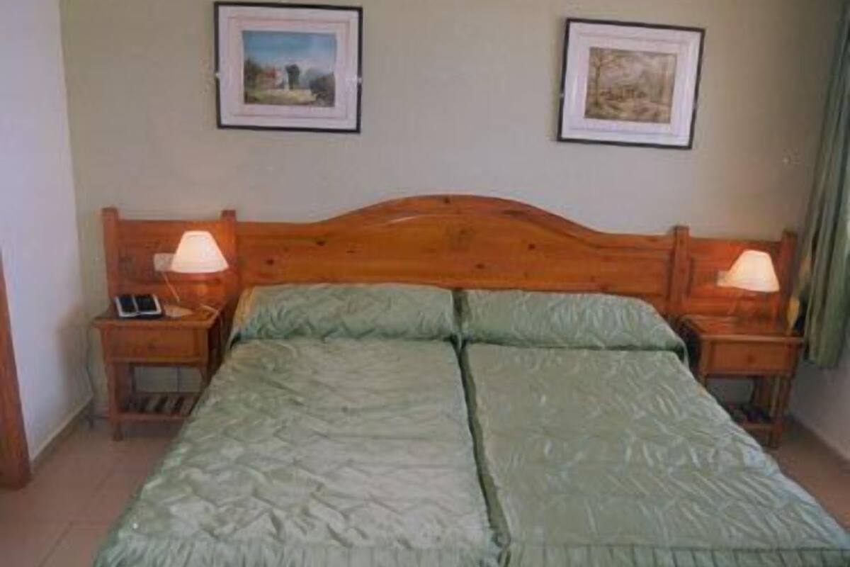 Una cama doble con colcha verde, dos mesitas de noche con lámparas, y dos cuadros enmarcados en la pared.