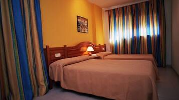 Habitación de hotel con dos camas, cabecera de madera, mesita de noche con lámpara y cortinas a rayas.