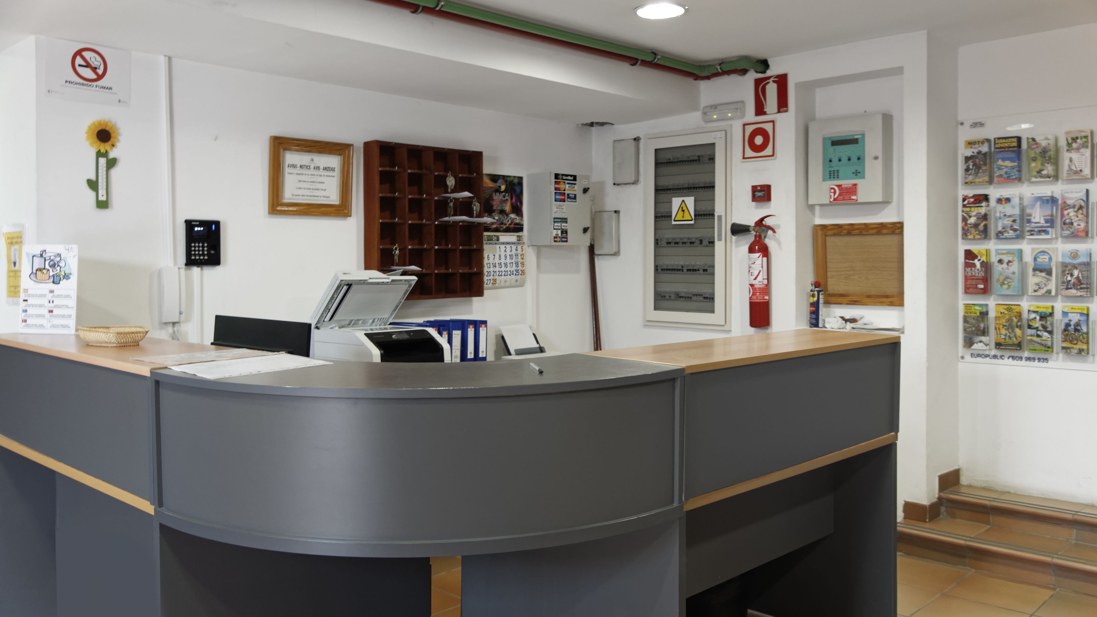 Un mostrador de recepción con una computadora, un extintor y una pared con varios carteles y un calendario.