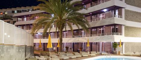A swimming pool with lounge chairs and umbrellas, surrounded by a building with multiple balconies and a palm tree.