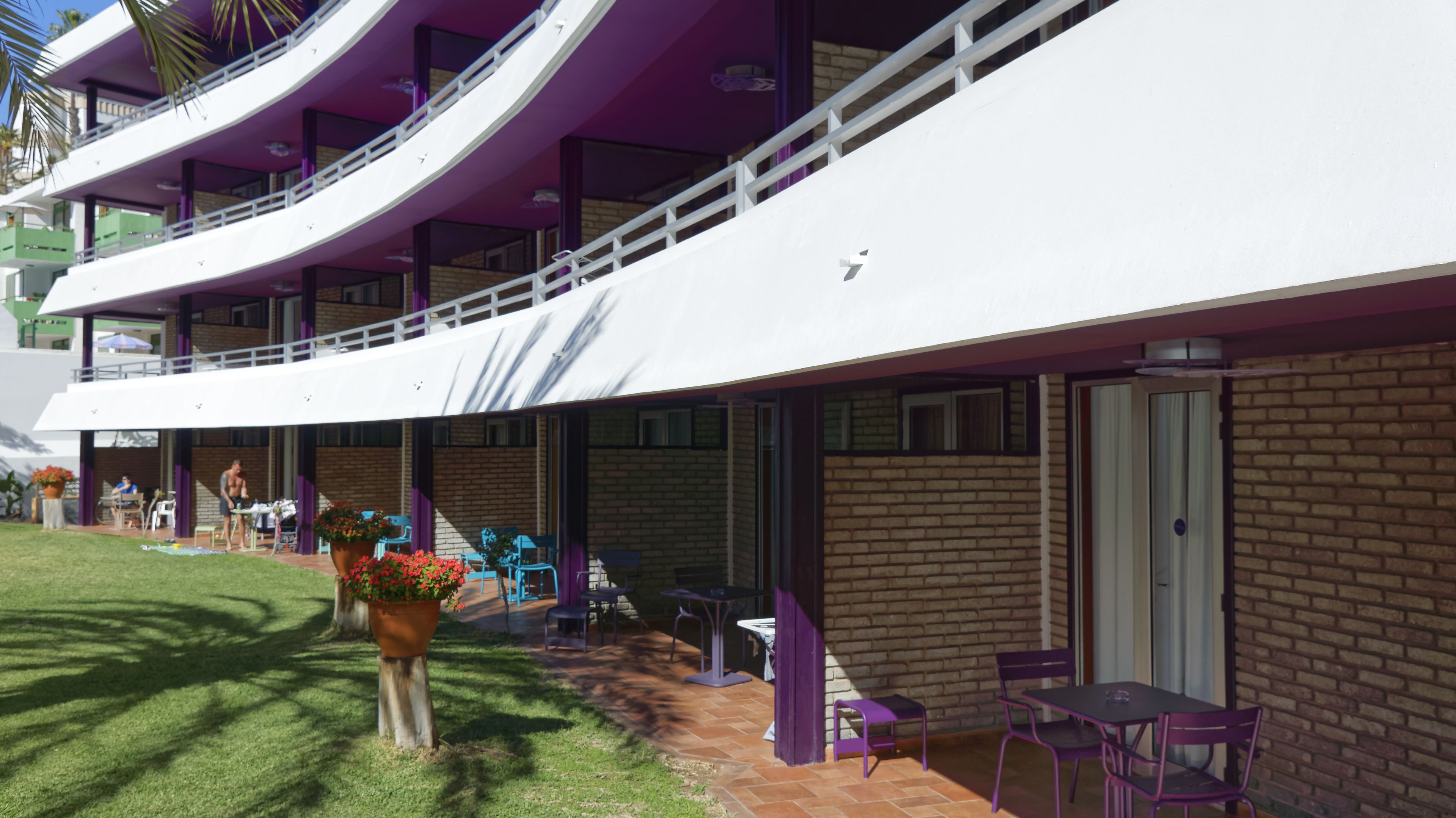 Un bâtiment moderne doté d’un toit blanc incurvé et de touches de couleur violette. Il propose des espaces de détente en plein air avec des tables et des chaises, ainsi qu’un mur de briques sur l’un de ses côtés.