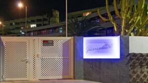 A gated entrance with a security sign and a neon sign for 'Jucarandinas Beach'. - Jacarandas Beach (San Bartolomé de Tirajana)