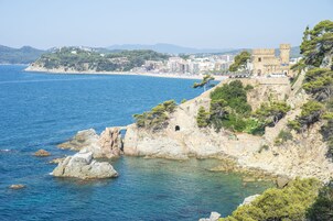 A coastal view with a castle on a cliff overlooking the sea, a beach, and a cityscape in the distance.