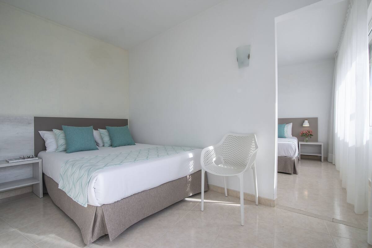 A modern bedroom with a bed, a white chair, a nightstand, and a small table with a lamp and flowers.