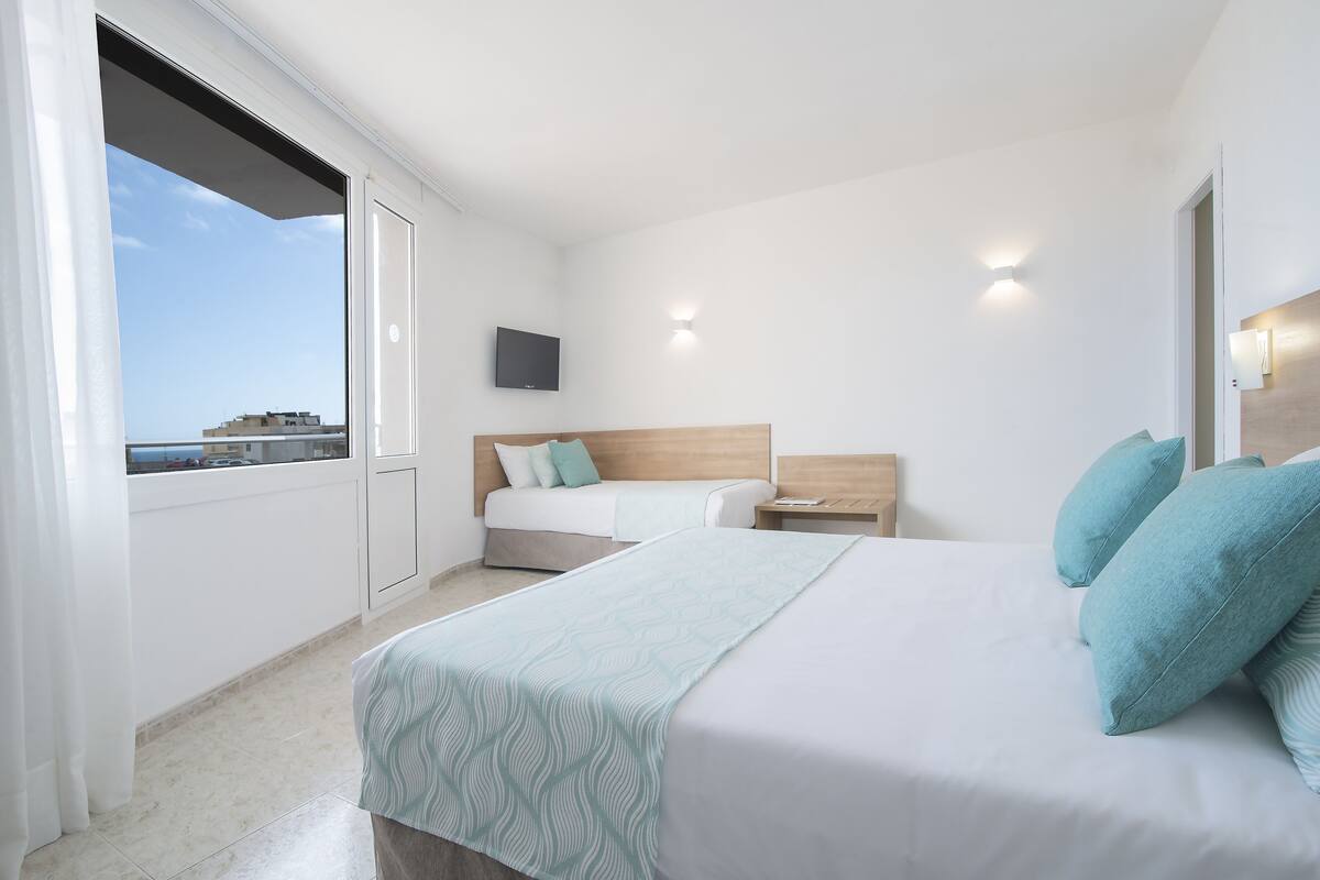 A hotel room with a bed, a bedside table, a TV mounted on the wall, and a window with a view of buildings.