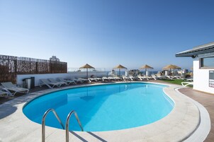 A swimming pool with sun loungers and umbrellas, surrounded by a white building and a wooden fence.