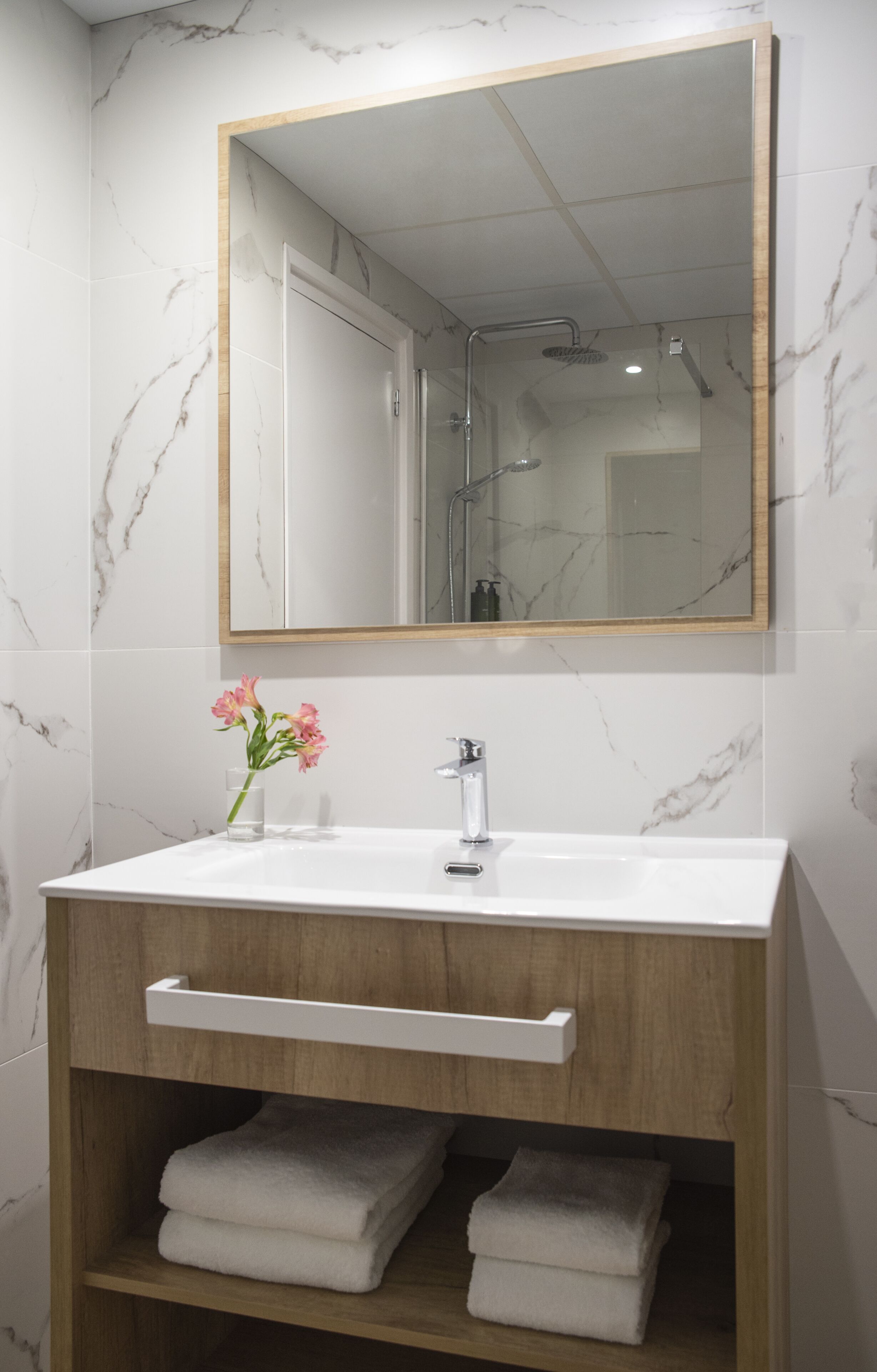 A bathroom with a marble-tiled wall, a wooden vanity with a white sink, a mirror, and a towel shelf.