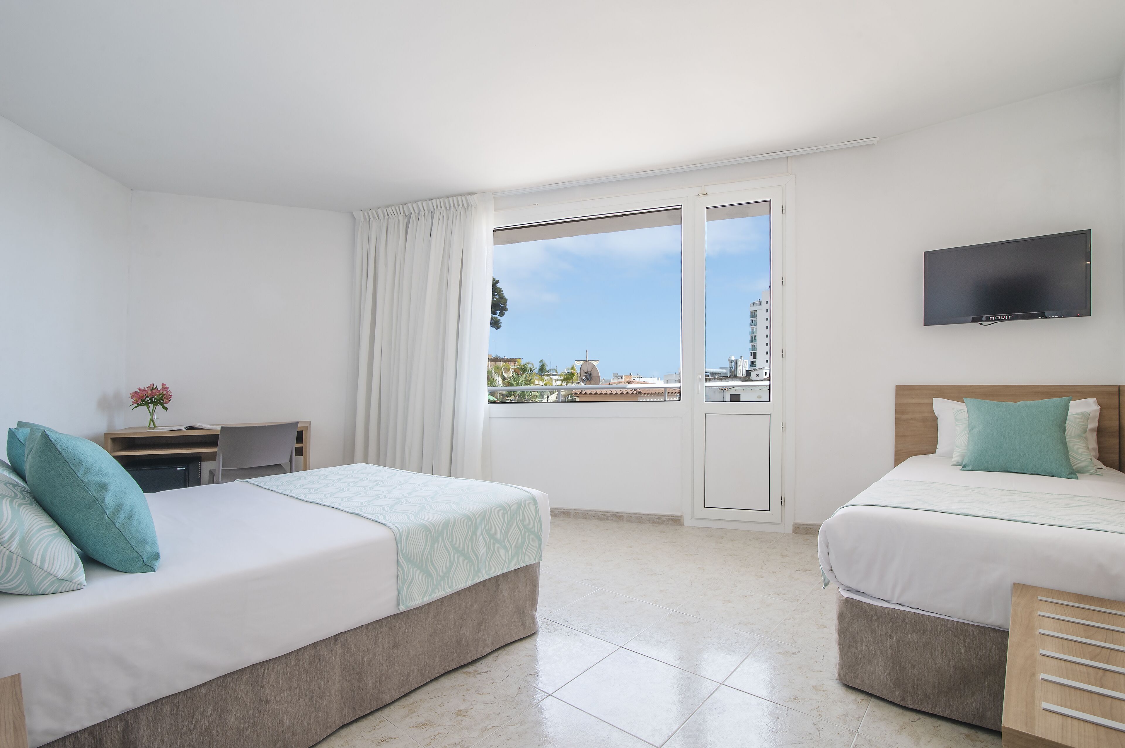 A hotel room with two beds, a desk, a TV, and a balcony with a city view.