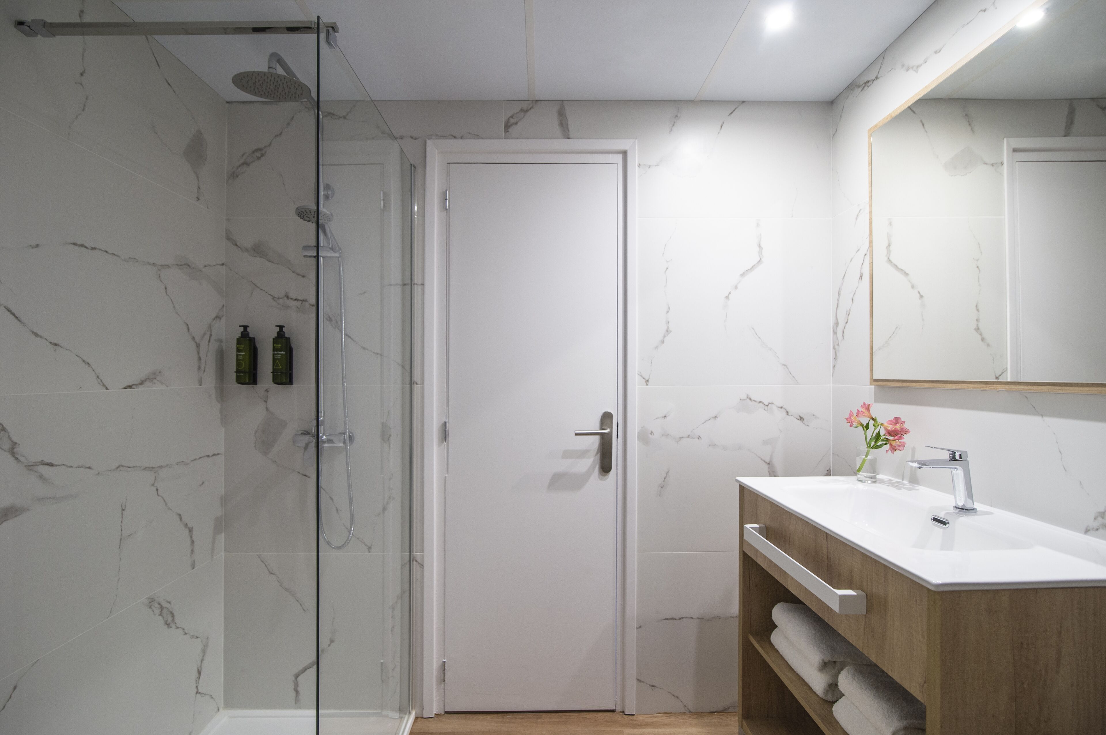 Un baño moderno con cabina de ducha de vidrio, una puerta blanca, un mueble de tocador de madera con lavabo y un revestimiento de pared de mármol.