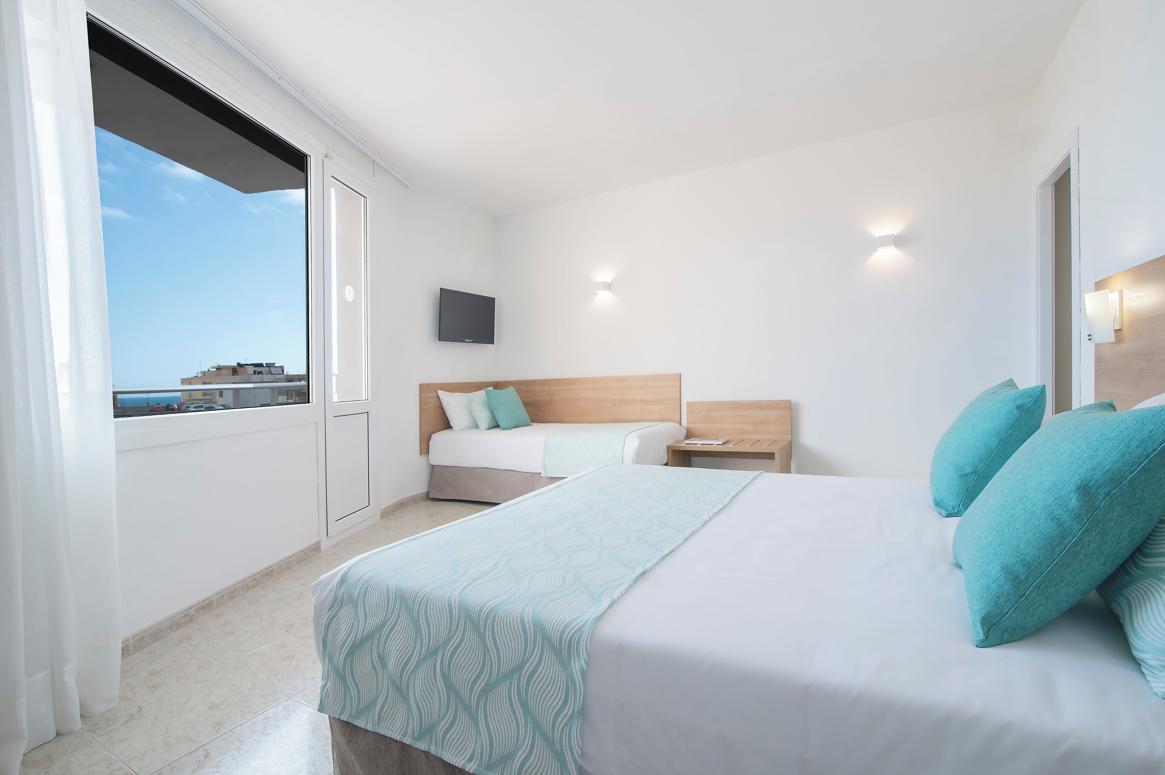 A hotel room with a bed, a bedside table, a TV mounted on the wall, and a window with a view of buildings.