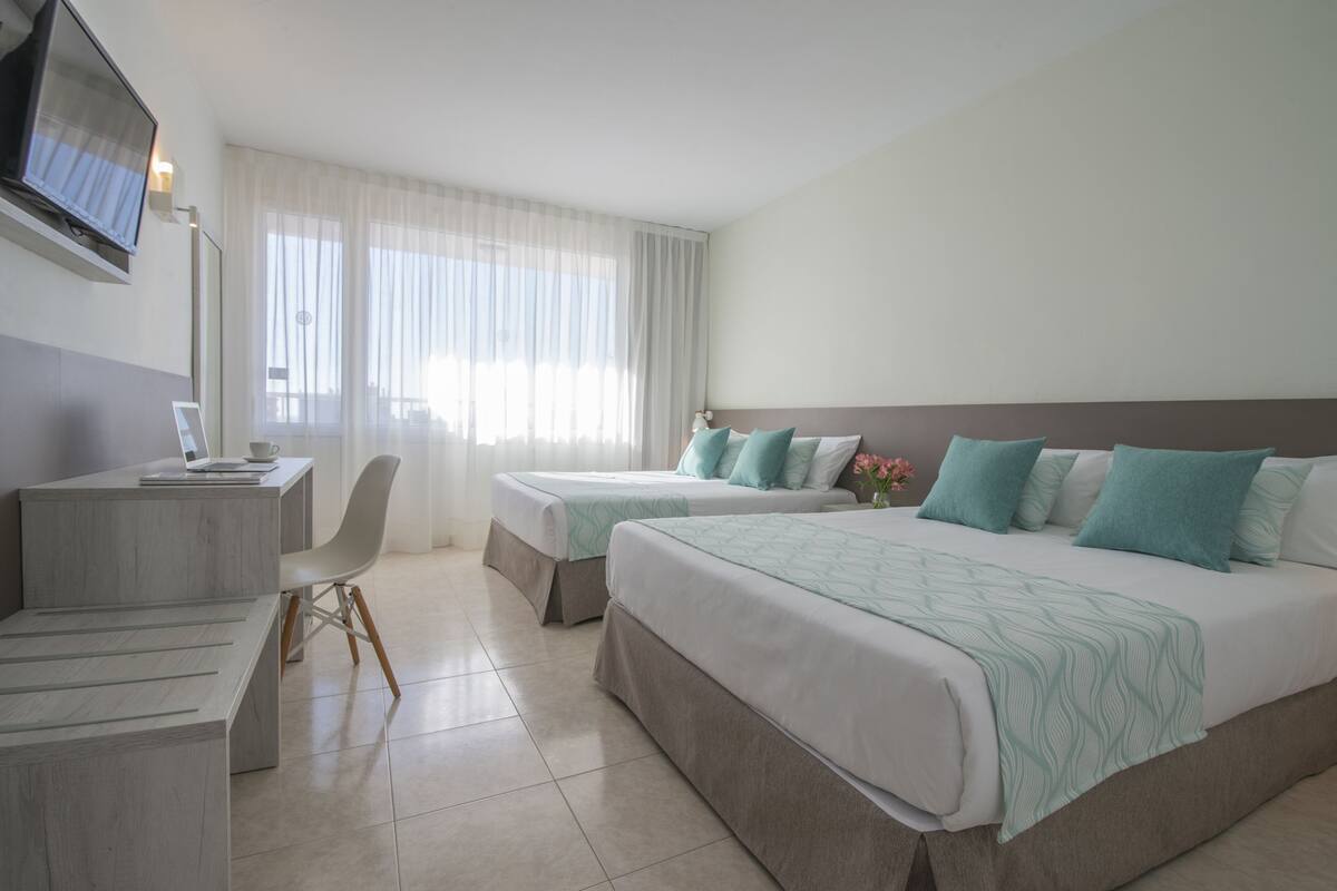 A hotel room with two beds, a desk with a chair, a TV, and a large window with sheer curtains.