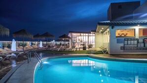 A swimming pool with lounge chairs and umbrellas, a building with a glass facade, and a thatched-roof structure in the background. - Hotel Lloret Santa Rosa by Pierre & Vacances (Lloret de Mar)