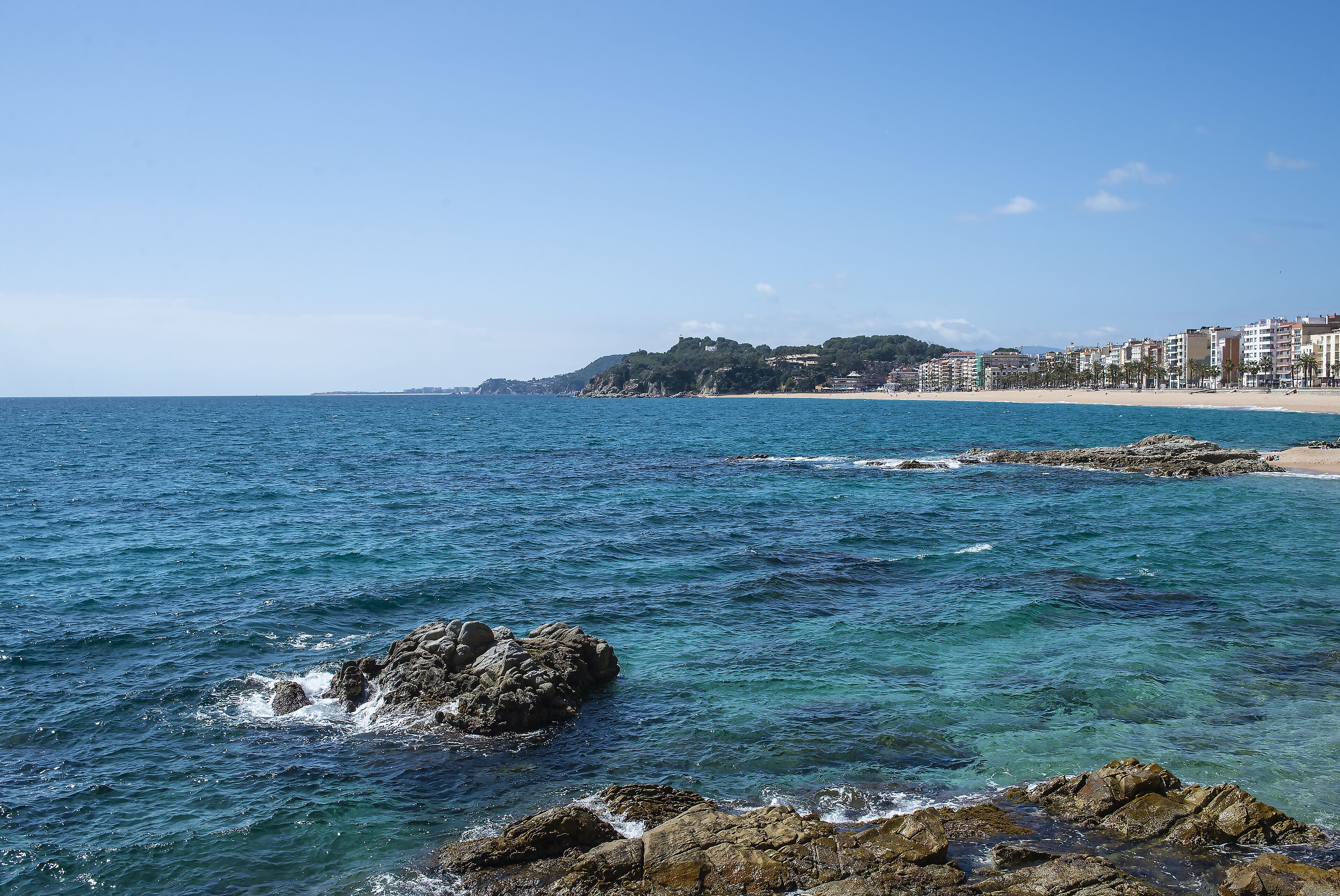 A coastal view with clear blue waters, rocky shoreline, and a sandy beach.