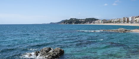 A coastal view with clear blue waters, rocky shoreline, and a sandy beach.