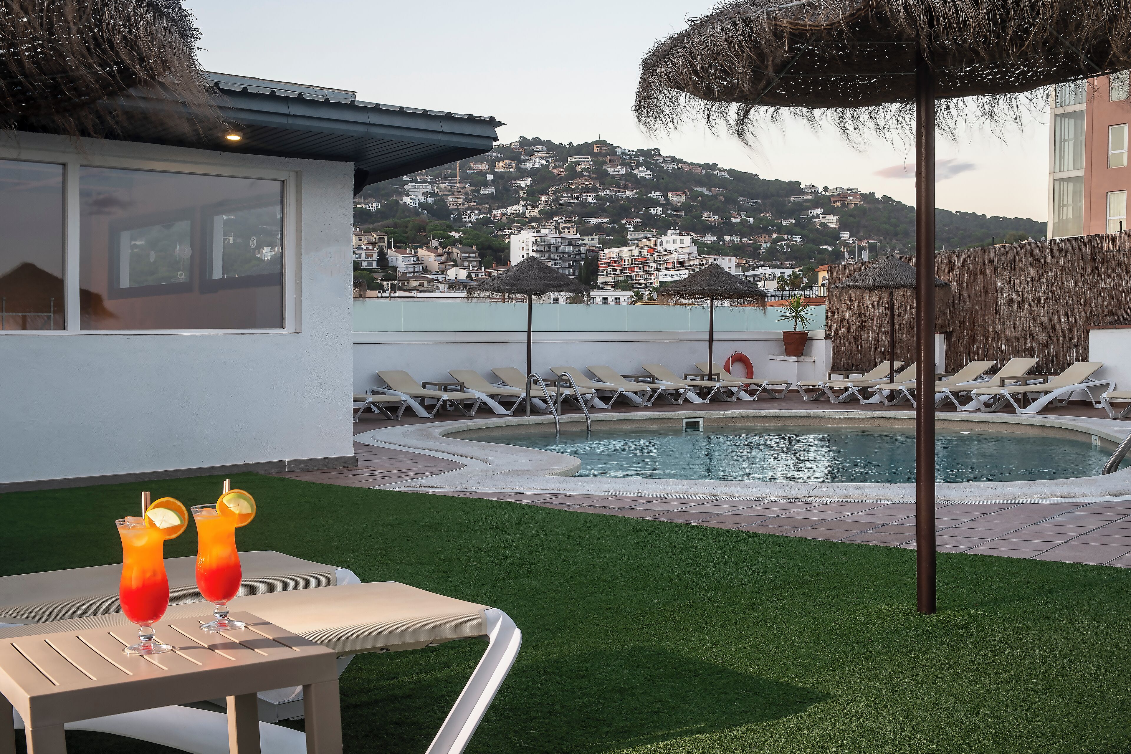 A poolside area with lounge chairs, a table with drinks, and a view of a hillside town.