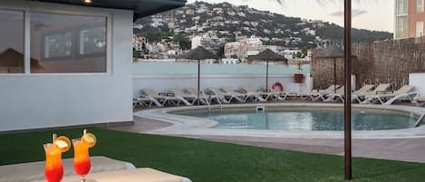 A poolside area with lounge chairs, a table with drinks, and a view of a hillside town.