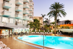 Una piscina con sillones de descanso y sombrillas, rodeada de palmeras y un edificio con balcones.