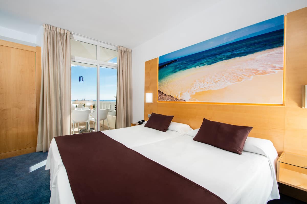 Une chambre d’hôtel avec un grand lit, un bureau avec une chaise, une cloison en verre et un tableau représentant une scène de plage.