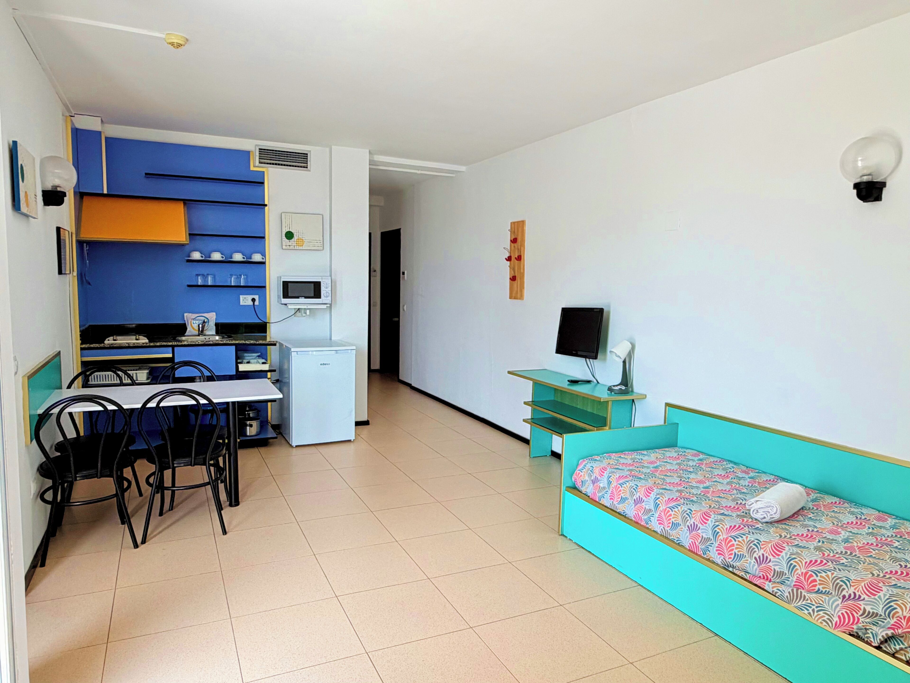 Habitación compacta con kitchenette, comedor, cama y escritorio con computadora.