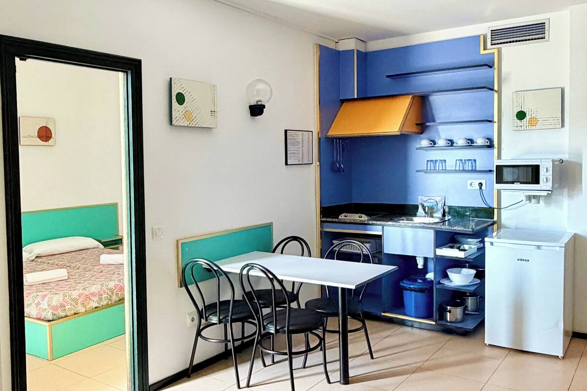A compact room with a dining table, chairs, a kitchenette, and a bed with a green headboard.