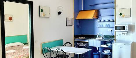 A compact room with a dining table, chairs, a kitchenette, and a bed with a green headboard.