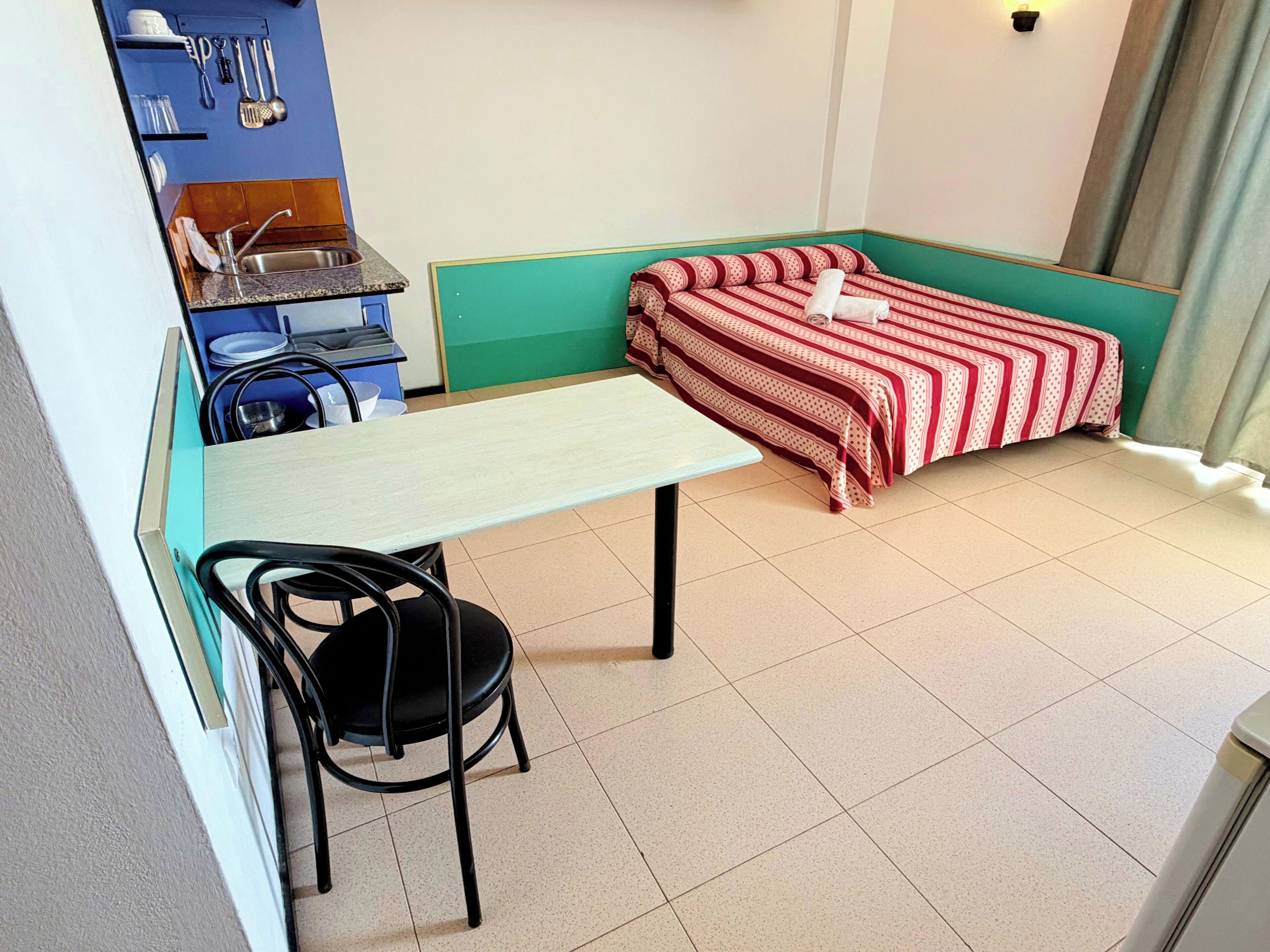 Habitación pequeña con cama, mesa, silla, lavamanos y estante con utensilios de cocina.