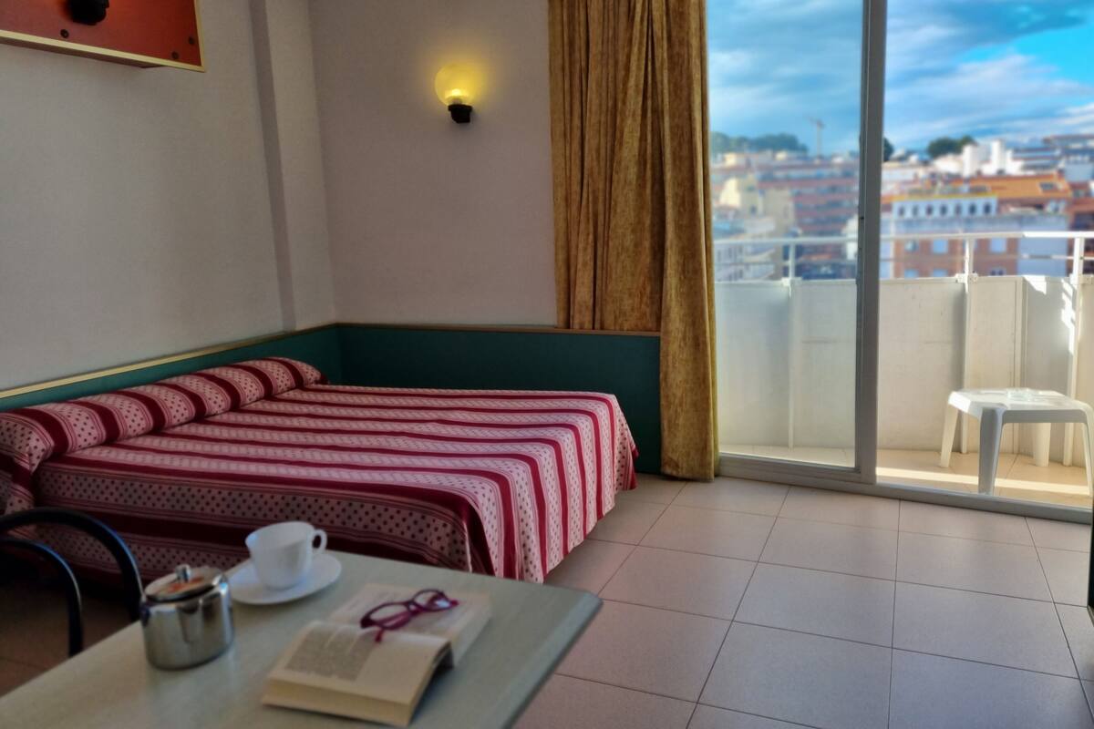 A hotel room with a bed, a table with a cup and book, and a view of the city through a large window.