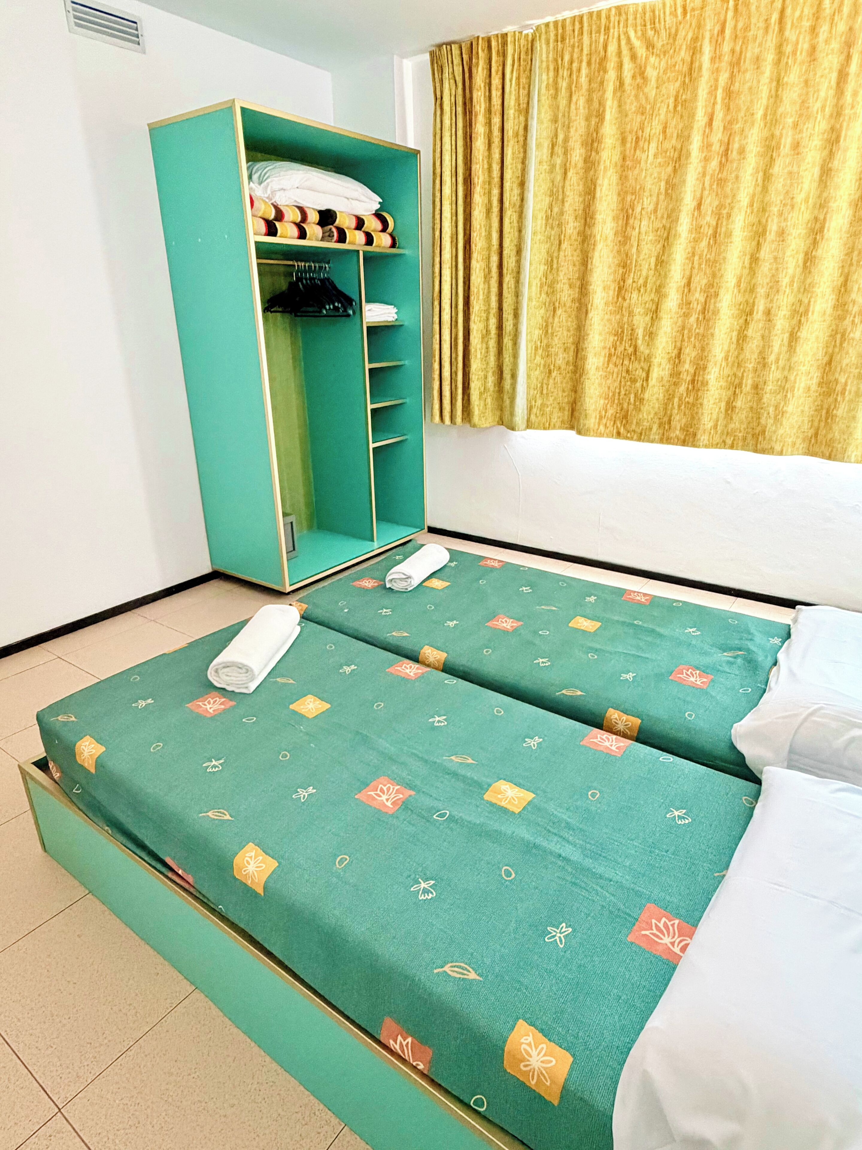 Una habitación con dos camas, un armario verde y una ventana con cortinas.