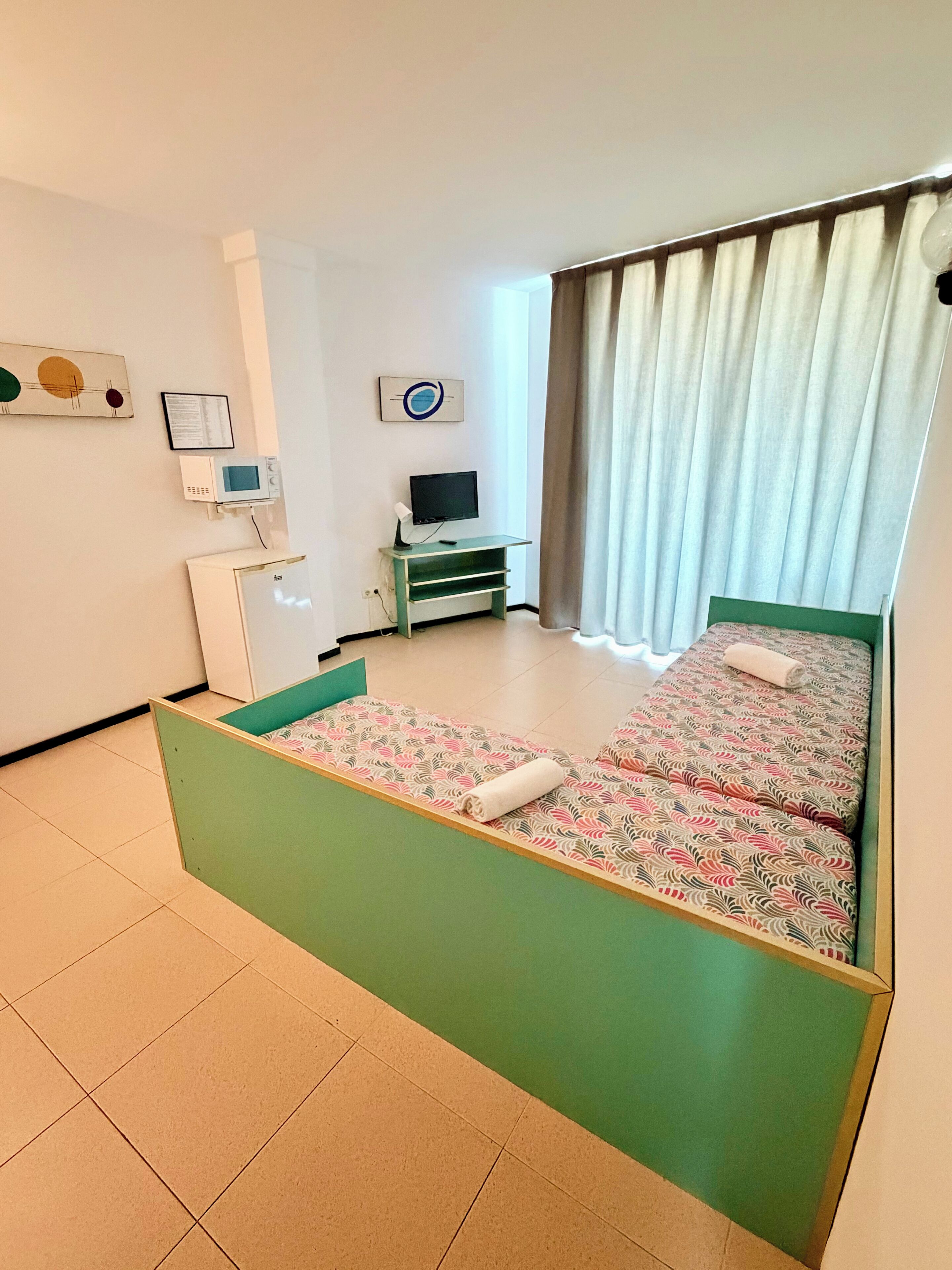 Quarto de hotel com cama, escrivaninha com computador, televisão e janela com cortinas.