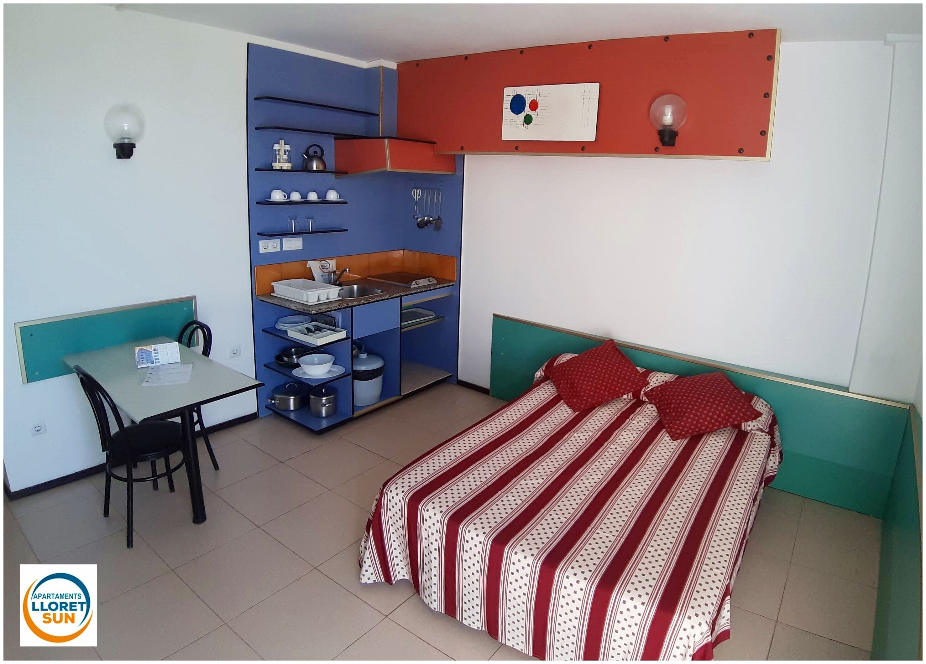 A compact bedroom with a bed, a small desk, and a kitchenette.