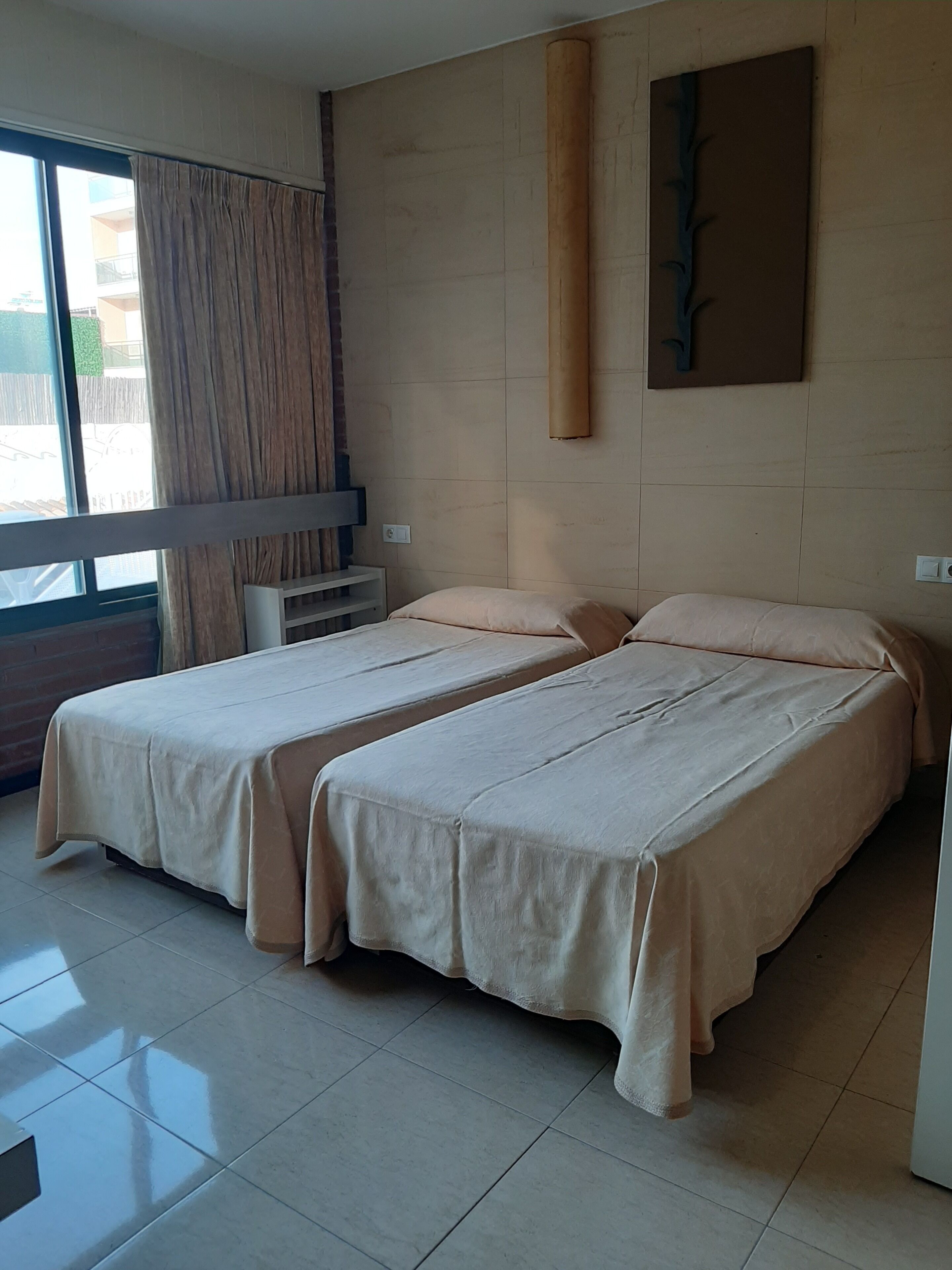 A hotel room with two beds, a large window with curtains, a wall-mounted air conditioner, and a plain wall with a decorative piece.
