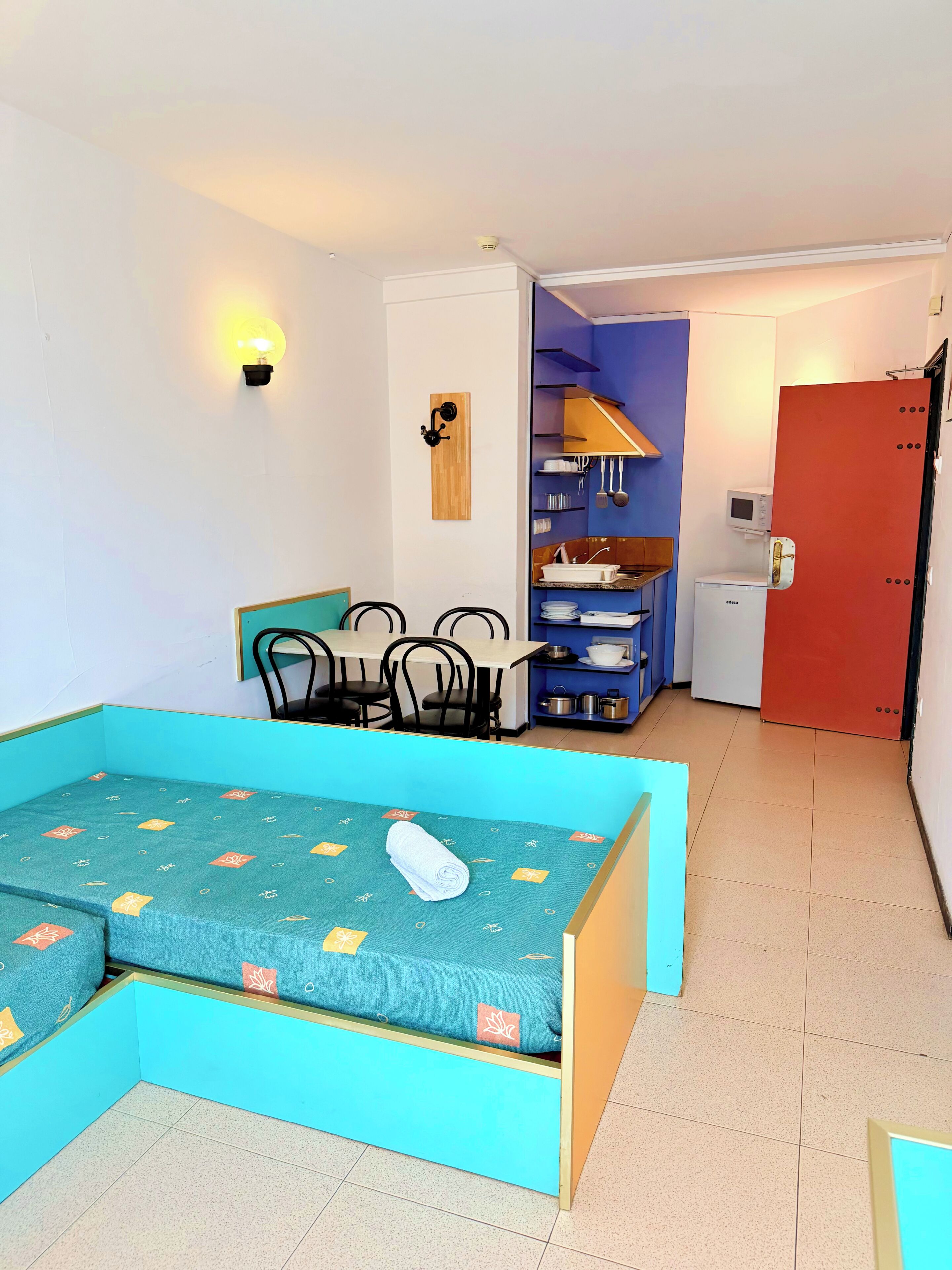Une pièce de taille modeste comprenant un lit, une petite table à manger avec des chaises, un coin cuisine et une porte rouge.