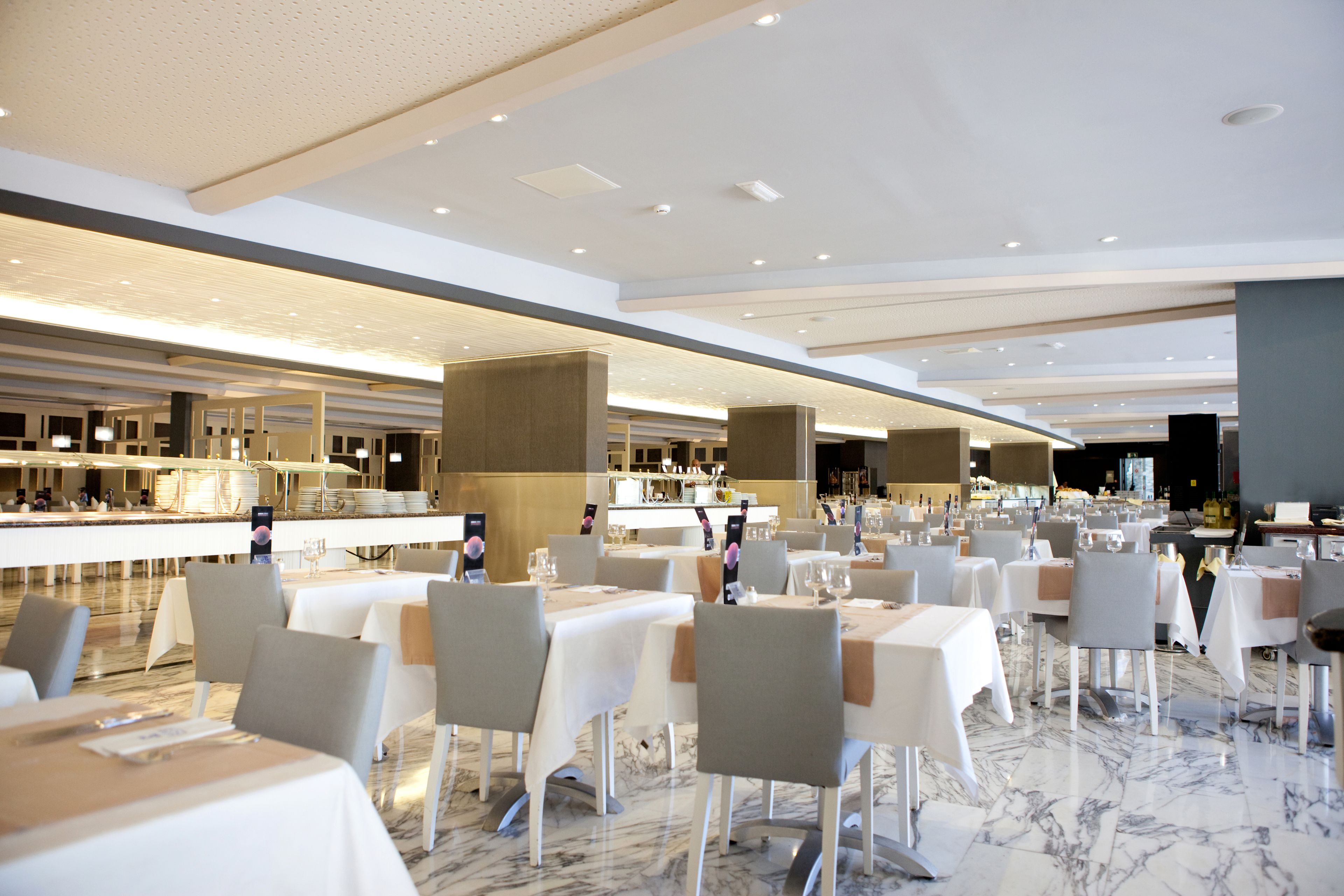 A spacious restaurant with multiple tables and chairs, a marble floor, and a modern ceiling design.